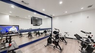 Located at 4410 Westheimer Rd in Houston, TX, this purpose-built exercise room serves as a dedicated indoor cycling studio. The space is thoughtfully designed to maximize functionality and motivation for users. A large, navy-blue-framed floor-to-ceiling mirror spans one full wall, visually expanding the room while allowing cyclists to monitor their form during workouts. A wall-mounted flat-screen television is positioned prominently above the mirror for entertainment or guided class viewing. The room accommodates multiple stationary spin bikes arranged to allow comfortable spacing between users, including StairMaster-brand cycles. Lighting is provided by recessed ceiling fixtures that illuminate the space evenly. The flooring consists of light wood-style planks that are both durable and easy to maintain. One accent wall features a striking white chevron-patterned tile, adding a modern, upscale design element to the room. Entry is through a glass-paneled door, and the room includes a hand sanitizer or wipe dispenser, a small waste receptacle, and posted yoga and facility policies. The clean white walls and crisp navy accents give the studio a polished, professional aesthetic well-suited for an urban Houston fitness facility.