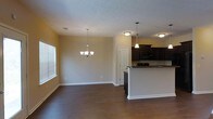This two-story home presents an open-concept main level where the living, dining, and kitchen areas flow seamlessly together. Dark hardwood flooring runs throughout the space, providing a warm and grounded aesthetic that contrasts with the neutral, light-toned walls and white trim. The dining area is anchored by an elegant multi-arm chandelier, while the kitchen features a pendant light above the breakfast bar peninsula, creating distinct lighting zones within the open layout.

The kitchen is outfitted with rich, dark wood cabinetry that extends to the upper walls, offering generous storage. A raised breakfast bar peninsula serves as a natural dividing element between the kitchen and the living area, making it ideal for casual dining or entertaining. Black appliances, including a built-in microwave and range, complement the dark cabinetry. A textured ceiling adds subtle dimension throughout the main level.

Natural light filters in through a window fitted with horizontal blinds, brightening the living space without compromising privacy. The overall ambiance is clean, functional, and cohesive, with a consistent design palette carried throughout the main level.