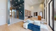 Located in Houston, TX, this single-story space presents an impressive communal environment defined by its dramatic architectural scale and contemporary design. Soaring double-height ceilings create an immediate sense of grandeur throughout, enhanced by an abundance of natural light streaming through floor-to-ceiling windows and striking arched glass panels that frame views of a lush outdoor pool area and surrounding landscaped grounds.

The main lounge area features light hardwood flooring throughout, anchored by generously sized neutral sectional sofas accented with blue and teal throw pillows. A bold teal-painted accent wall serves as a focal point, hosting a wall-mounted flat-screen television. Decorative wall art, including a cluster of sculptural circular pieces in blue tones, adds artistic character to the space.

An open reception or lobby area features a built-in light wood shelving unit that spans an entire wall, providing both functional storage and a striking visual element. A dark teal structural column and bold contemporary artwork contribute to the modern aesthetic. Recessed lighting and large black-framed windows further complement the clean, sophisticated interior throughout this well-appointed space.
