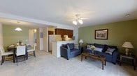 This single-floor residence offers a welcoming open-concept design that connects the living, dining, and kitchen areas into one cohesive, functional space. The living area is carpeted in light neutral tones, creating a warm and comfortable atmosphere, and is accented by a sage green feature wall that adds a touch of color and character. A ceiling fan with integrated lighting serves as the central fixture overhead, providing both style and practicality. The living space comfortably accommodates a sofa and loveseat set in a deep navy blue, complemented by a rustic wood-top coffee table that grounds the room. The kitchen is positioned adjacent to the living area and is partially separated by a raised breakfast bar counter, maintaining an open feel while defining the cooking space. Rich wood-toned cabinetry lines the upper walls of the kitchen, and a white refrigerator and countertop appliances complete the setup. The layout also features a connected dining area, visible to the side, with bright white walls and tiled flooring that distinguish it from the carpeted living zone. Overall, the space presents a clean, practical, and cozy environment suited for comfortable everyday living.