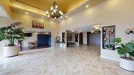 This single-floor lobby presents a polished and welcoming environment designed to make a strong first impression. The entrance features floor-to-ceiling glass panels flanking a set of sleek double doors, flooding the foyer with natural light and offering clear views of the surrounding landscape. A herringbone-patterned marble-style tile floor extends throughout, lending a sense of refinement to the space.

The central lobby is anchored by a statement chandelier suspended from a raised, gold-toned ceiling tray, adding warmth and visual drama overhead. Recessed lighting complements the chandelier, ensuring the space remains well-illuminated throughout. The open layout is generously proportioned, allowing for comfortable movement across the area.

A dedicated reception area features a dark wood-paneled desk with a granite countertop, framed by a large window that brings in additional natural light. Nearby, a lounge alcove includes bar-height seating, a wall-mounted television, and built-in package lockers, offering practical amenities within the lobby environment.

The elevator bank is situated at the rear, flanked by decorative niches, wall art, and cushioned seating, creating a refined waiting area. Curated artwork, potted plants, and accent lighting are distributed throughout, contributing to the space's warm, sophisticated ambiance.