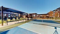 This expansive outdoor amenity space is thoughtfully designed to serve as a central hub for recreation and relaxation within a modern multi-story residential community. At the heart of the space are multiple pickleball courts with vibrant blue and green court surfaces, complete with regulation nets, offering residents a dedicated area for both casual play and competitive matches.

Adjacent to the courts, large shade canopies provide comfortable seating areas furnished with Adirondack-style and slatted chairs, creating inviting spots to relax before or after activity. The hardscaped areas transition seamlessly into manicured green lawns, while carefully arranged ornamental plantings, decorative rock beds, and young trees add texture and visual interest throughout the grounds.

Winding concrete pathways connect the various zones of the amenity area, encouraging leisurely strolls through the landscaped environment. Additional pergola-style shade structures are positioned throughout, extending the usability of the outdoor space across varying weather conditions. The surrounding residential buildings feature a contemporary architectural design with a mix of brick, vertical board-and-batten siding, and large balconies. Under open skies, the entire amenity area is bathed in abundant natural light, creating a bright, welcoming atmosphere that enhances the overall outdoor living experience.