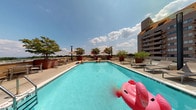 This amenity space is situated at pool level within a multi-story residential building, offering convenient and direct access to a well-appointed outdoor pool deck. The interior transitional areas feature neutral-toned carpeted flooring and clean white walls, with glass-paneled doors that draw in natural light and provide clear sightlines to the outdoors. The pool deck itself is generously sized, featuring a large rectangular swimming pool with clear turquoise water, surrounded by a broad concrete surround outfitted with lounge chairs, shaded umbrella stations, and potted trees that add greenery to the space. The deck is enclosed by a low railing that opens up expansive views of the surrounding urban landscape and neighboring buildings. A sleek, modern aesthetic is maintained throughout the outdoor area, with coordinated furnishings including chaise lounges and casual seating arrangements suited for relaxation. The building's bold, multi-story façade of warm brown tones and stacked balconies forms a striking backdrop to the pool area, reinforcing the urban character of the environment. Overall, the space creates a welcoming and functional outdoor retreat within a dense city setting.