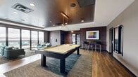 Located in Centennial, CO, the Sky Deck is a generously sized single-floor rooftop amenity space designed for socializing and relaxation. The interior is thoughtfully divided into several distinct zones, each with its own character and purpose. A game room area anchors one section of the space, highlighted by a sleek billiards table beneath a modern linear pendant light, a wall-mounted TV, decorative geometric mirrors, and bar-height seating along an exposed brick accent wall. Adjacent to this is a lounge area featuring a bold, floor-to-ceiling abstract striped wallcovering, complemented by teal and neutral sofas arranged around a wood coffee table. Another seating nook showcases plush golden armchairs positioned to take full advantage of floor-to-ceiling sliding glass doors that flood the room with natural light and open directly onto the outdoor deck. The outdoor terrace, visible through expansive glazing, extends the living space further with additional seating. Wide-plank hardwood-style flooring flows throughout, tying the zones together cohesively. The varied ceiling treatments, including tray ceilings with recessed lighting and a rich dark-finished ceiling in the game room, add architectural depth. The overall ambiance is contemporary and inviting, offering a refined space for both active recreation and leisurely gathering.
