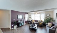The Kennedy is a thoughtfully designed 2-bedroom, 2-bathroom floor plan laid out entirely on a single level for convenient, accessible living. The living area immediately captures attention with its bold accent wall in a deep plum tone, creating a striking contrast against the neutral ceiling and light-toned walls. Rich, wood-style hard-surface flooring runs throughout, adding warmth and visual continuity to the space. A textured ceiling adds subtle dimension overhead, while a ceiling fan with a light fixture ensures year-round comfort. Multiple windows dressed in sheer white curtains invite generous natural light, creating a bright and airy atmosphere. The living room offers ample space for comfortable seating arrangements, with room for both a media console and a central coffee table. The overall ambiance strikes a balance between bold design choices and a welcoming, livable aesthetic. The two bedrooms and two full bathrooms provide practical separation of space, making the Kennedy an ideal layout for roommates, guests, or those who prefer a dedicated home office. With its combination of contemporary finishes and functional design, the Kennedy floor plan delivers a comfortable and stylish living experience.