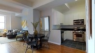 This single-floor space showcases an open-concept layout that seamlessly connects the dining area and kitchen, creating a cohesive and functional living environment. The dining area features warm hardwood flooring and is furnished with a round dark-finished table surrounded by classic Windsor-style chairs. A decorative mirror leans against the wall, amplifying natural light and adding depth to the space, while a vibrant yellow abstract artwork and a tall vase with branching greenery contribute a stylish, curated aesthetic.

The kitchen transitions naturally from the dining area, marked by a shift to dark tile flooring. It features white upper cabinetry that extends to the ceiling, providing generous storage, contrasted by dark countertops that lend a modern touch. A stainless steel sink sits beneath a clean backsplash, and stainless steel appliances, including a built-in dishwasher and microwave, complete the kitchen's contemporary look. The layout is efficiently organized, offering ample counter space for meal preparation.

In the background, a glimpse of a living area with a sofa and accent cushions adds to the sense of a well-connected, multi-functional living space with a warm and inviting ambiance throughout.