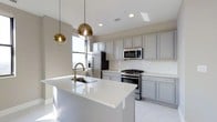 Located at 50 W Broad St in the heart of Columbus, Ohio, this single-floor space offers a refined, contemporary aesthetic highlighted by a thoughtfully designed kitchen. The kitchen serves as a central feature, showcasing soft gray cabinetry that extends to the ceiling, providing generous storage while maintaining a clean, cohesive look. A large central island with a bright white quartz countertop anchors the space, equipped with an undermount sink and a brushed gold faucet that adds a warm, modern accent. Overhead, two globe pendant lights in a gradient gold finish hang elegantly above the island, contributing both task lighting and visual interest. Stainless steel appliances, including a gas range, built-in microwave, and refrigerator, complement the kitchen's modern character. Recessed lighting throughout ensures the space is well-lit, while a window allows natural light to fill the area. The light-toned flooring ties the room together, reinforcing the bright, open ambiance. The overall design balances functionality with sophisticated style, making it a polished and practical space in a prime downtown location.