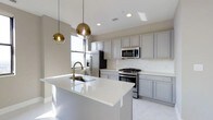 Located at 50 W Broad St in the heart of Columbus, Ohio, this single-floor space offers a refined, contemporary aesthetic highlighted by a thoughtfully designed kitchen. The kitchen serves as a central feature, showcasing soft gray cabinetry that extends to the ceiling, providing generous storage while maintaining a clean, cohesive look. A large central island with a bright white quartz countertop anchors the space, equipped with an undermount sink and a brushed gold faucet that adds a warm, modern accent. Overhead, two globe pendant lights in a gradient gold finish hang elegantly above the island, contributing both task lighting and visual interest. Stainless steel appliances, including a gas range, built-in microwave, and refrigerator, complement the kitchen's modern character. Recessed lighting throughout ensures the space is well-lit, while a window allows natural light to fill the area. The light-toned flooring ties the room together, reinforcing the bright, open ambiance. The overall design balances functionality with sophisticated style, making it a polished and practical space in a prime downtown location.