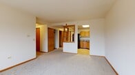 Located at 3 Lilac Ave in Fox Lake, Illinois, this one-bedroom, one-bathroom unit offers a practical and well-organized layout across a single floor. The combined kitchen and living area forms the heart of the unit, where a half-wall with open wood shelving visually separates the two spaces while maintaining an open feel. The kitchen features warm oak cabinetry, granite-patterned countertops, a stainless steel sink, and a full-size refrigerator, with light hardwood-style flooring underfoot. The living area is carpeted and bathed in natural light from a multi-pane window, creating a comfortable and inviting atmosphere. Baseboard heating runs throughout the unit, ensuring consistent warmth. The bedroom is a well-proportioned, carpeted retreat with a large sliding-door closet offering ample storage and a window with natural views of the surrounding landscape. Two hallways connect the main living spaces to the bedroom and bathroom, providing a sensible traffic flow. The bathroom includes a full soaking tub with shower, a wall-mounted vanity sink, tile flooring, and a mirrored medicine cabinet. Neutral tones and warm wood accents throughout lend the unit a cohesive, understated character.