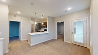 This well-appointed multi-level residence is designed with a practical, modern layout that balances open communal spaces with functional private areas. The kitchen serves as a central highlight, featuring dark espresso cabinetry, stainless steel appliances including a range, microwave, and dishwasher, and a white countertop peninsula with pendant lighting overhead. Light wood-look flooring grounds the kitchen, which opens directly to the main living area. The living space is finished with neutral carpet and benefits from natural light through a glass exterior door. A carpeted staircase with a metal handrail connects to the upper level, where a bright, open loft area offers flexible use of space, illuminated by recessed lighting and windows with exterior views. The upper landing provides access to additional rooms, including a carpeted bedroom with a ceiling fan and window, a walk-in closet outfitted with wire shelving systems, and a full bathroom featuring dark vanity cabinetry, a white countertop, and a tub-shower combination. A dedicated laundry room accommodates a stacked washer and dryer unit alongside a water heater, with easy-care wood-look flooring throughout.