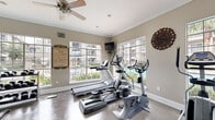 Located in Houston, TX, this fitness center is a well-equipped exercise space designed to support a complete workout routine. The room is finished with a durable speckled floor, crisp white crown molding, and neutral-toned walls that create a clean, functional atmosphere. Large multi-pane windows line the walls, flooding the space with abundant natural light while offering views of an outdoor pool and surrounding greenery. A ceiling fan overhead provides additional air circulation throughout the room.

The cardio area features a treadmill, elliptical trainer, and stationary bike, all positioned to take advantage of the bright, outward-facing windows. On the strength training side, the space includes a cable machine, a weight bench, and a rack of dumbbells, offering a range of resistance options. Full-length mirrors span one wall, visually expanding the space and allowing users to monitor their form during workouts. A wall-mounted flat-screen television is also present, providing entertainment during exercise sessions.

A set of elegant French doors with transom windows serves as the primary entrance, adding an architectural touch that elevates the overall aesthetic. The fitness center rules are prominently displayed near the entrance, and decorative wall accents add a subtle design element. Overall, this exercise room delivers a bright, practical, and well-rounded fitness environment.