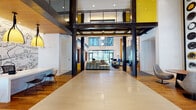 This expansive lobby makes an immediate impression with its soaring double-height ceilings, bold architectural framework of exposed dark steel columns, and a vibrant color palette anchored by striking yellow accents. The entry corridor features large-format polished tile flooring that leads the eye toward floor-to-ceiling glass doors and windows, flooding the space with abundant natural light and views of the surrounding streetscape.

The reception area is anchored by a sleek, minimalist white desk illuminated by a trio of pendant lights with warm yellow shades, set against a dramatic hand-drawn illustrative mural that adds a playful, artistic character to the space. Warm hardwood flooring defines the lounge zones, where generously sized sectional sofas in multicolored patterned upholstery are arranged around coffee tables and accented with blue lounge chairs.

A standout feature is the tall, backlit shelving wall clad in warm wood tones, displaying a colorful arrangement of books and decorative objects alongside a substantial stone fireplace that anchors the lounge. Additional seating nooks framed in rich walnut with yellow-backed panels provide intimate gathering spots. Sculptural pendant fixtures and carefully layered lighting throughout complete the refined, design-forward ambiance of this welcoming communal space.
