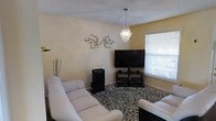 This single-floor space offers a welcoming and thoughtfully designed interior with a distinct sense of personality and style. The living area serves as the centerpiece of the home, featuring white upholstered seating arranged around a patterned area rug that anchors the room with a bold, classic motif. A striking crystal chandelier hangs from the textured ceiling, lending an elegant focal point that contrasts beautifully with the casual comfort of the seating arrangement. Natural light filters in through a window fitted with blinds, brightening the warm, neutral-toned walls. Decorative wall art in the form of a branching floral metal piece adds an artistic touch, contributing to the room's curated ambiance. An entertainment setup, complete with a large flat-screen television, media stand, and audio equipment, provides a fully functional and modern media experience. The combination of refined decorative elements and practical furnishings creates a space that balances elegance with everyday livability, making it both an inviting and functional environment.