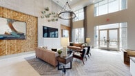 Located at 1401 Dixie Hwy in Covington, Kentucky, this elegantly designed amenity space offers a refined and welcoming environment that blends modern aesthetics with comfortable functionality. The centerpiece of the space is a dramatic double-height lounge area, where soaring ceilings amplify the sense of openness and grandeur. A striking circular chandelier anchors the room, casting warm light over carefully curated seating arrangements that include plush sofas, tufted loveseats, and mid-century modern accent chairs arranged around a rustic wood coffee table. Textured wall panels — including woven and honeycomb-patterned surfaces — add warmth and visual depth, while decorative ceramic wall art introduces organic, artistic detail. Floor-to-ceiling windows flood the space with abundant natural light and frame views of the surrounding cityscape. An adjacent billiards area continues the double-height design language, featuring a full-size pool table surrounded by additional lounge seating and expansive glazed walls that reinforce the airy, open atmosphere. A separate kitchenette and coworking nook rounds out the space, outfitted with a long white quartz countertop, gray cabinetry, a coffee station, and a wall-mounted television backed by a striking reclaimed wood accent wall, providing a functional yet stylish setting for work or casual gatherings.