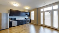 This single-floor space offers a thoughtfully designed open-concept layout that seamlessly blends the kitchen and living areas into one cohesive, functional environment. The kitchen is outfitted with sleek, dark cabinetry that extends to the upper walls, providing ample storage, and is complemented by light granite countertops that offer a striking contrast. Stainless steel appliances, including a gas range and dishwasher, along with a built-in microwave, complete the modern kitchen setup. The space flows effortlessly into the living area, where warm hardwood flooring stretches throughout, lending the interior a clean, contemporary feel. Natural light pours in generously through large windows fitted with blinds, as well as through a set of elegant French doors that brighten the entire space and create a welcoming, airy ambiance. The neutral-toned walls enhance the sense of openness, while flush-mount ceiling lights provide consistent illumination throughout. The overall design balances practicality with modern aesthetics, making this a versatile and inviting space suited for a variety of uses.