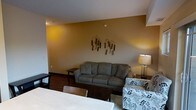 This single-floor space showcases a combined living and dining area that flows seamlessly together. The living area is anchored by a gray upholstered sofa and complemented by a floral-patterned accent chair, offering comfortable seating arrangements. A warm amber-toned accent wall serves as a focal point behind the sofa, adorned with decorative botanical wall art featuring cattail-inspired pieces that add a natural, earthy touch to the space. The walls are finished in a neutral tone that pairs well with the warm accent wall. A table lamp with a classic shade provides ambient lighting, supplementing the overhead lighting. Dark hardwood-style flooring runs throughout, adding warmth and continuity to the space. The dining area, partially visible in the foreground, features a light-colored table and wooden chairs, providing a functional space for meals. The overall design aesthetic combines neutral tones with warm accent colors, creating a cozy and welcoming ambiance. The open-plan layout between the living and dining areas enhances the sense of space and encourages a natural flow between functional zones.