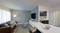 This single-floor space showcases a thoughtfully designed open-concept layout that brings together the living and kitchen areas in a cohesive, modern aesthetic. The living area is anchored by a striking large-format concrete-panel accent wall, which lends an industrial yet refined character to the space. A wall-mounted flat-screen television sits above a sleek white floating media console, creating a clean and contemporary focal point. The living area features light wood-toned flooring, a textured area rug, and a comfortable sofa accented with decorative pillows, complemented by a geometric side table that adds visual interest. A modern arc floor lamp provides warm ambient lighting. The open kitchen is accessible directly from the living space and is defined by dark espresso cabinetry paired with crisp white countertops, including a peninsula that serves as both a prep surface and a casual dining counter with seating. The kitchen is equipped with a stainless steel sink and cooking range, with the cabinetry providing ample storage. The contrast between the dark cabinetry and white surfaces creates a bold, polished look. The overall ambiance of the space is modern and urban, with a neutral palette of grays, whites, and dark tones that work harmoniously throughout.