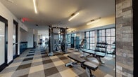 This contemporary fitness center is thoughtfully designed to accommodate a full range of workout routines across several distinct zones. The space features a striking patterned carpet tile floor in neutral tones of tan, gray, and brown, lending a modern and cohesive aesthetic throughout. Linear LED lighting runs across the ceilings, providing bright, even illumination that complements the natural light streaming in through large louvered windows along the exterior walls.

The main strength training area is generously outfitted with Matrix-brand cable machines, adjustable benches, and a comprehensive dumbbell rack, all arranged to allow ample space for movement. Mirrored wall panels enhance the sense of openness and support proper form during workouts. An adjacent cardio zone houses multiple treadmills, elliptical trainers, and stair climbers, all positioned to take advantage of outdoor views through expansive windows.

A dedicated cycling studio features stationary bikes with digital monitors, wall-mounted fans for ventilation, and additional louvered windows that bring in natural light. The entry area is distinguished by richly textured stone-look wall cladding in warm brown tones, creating a stylish transition into the facility. This vestibule also includes a water station and access to restrooms. The overall ambiance is polished, functional, and inviting, making this fitness center a well-rounded destination for exercise.