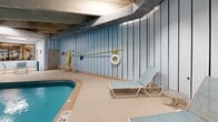 Located at 80 N Portage Path in Akron, Ohio, this expansive indoor recreational facility offers a comprehensive suite of amenities across a generously sized single floor. The centerpiece of the space is a large indoor swimming pool, framed by a distinctive exposed concrete beam ceiling that lends the area an industrial yet functional character. Light-blue vertical panel walls surround the pool deck, creating a cohesive and calming aesthetic throughout.

The pool area is well-equipped for safety and leisure, with poolside lounge chairs, a life ring station, and clearly posted safety signage. A separate hot tub is conveniently situated adjacent to the main pool, providing a dedicated relaxation zone. Casual seating, including a round table with chairs, offers a comfortable gathering area for swimmers and guests.

A glass-partitioned fitness center is directly accessible from the pool deck, housing a variety of strength and cardio equipment visible through large windows. The gym is well-lit with overhead fluorescent lighting, ensuring a bright and functional workout environment.

The facility also includes a staircase providing vertical access, along with additional utility and support rooms. The tile flooring throughout the pool deck is practical and durable, designed to handle a high-traffic aquatic environment. Overall, this facility is a well-appointed, multi-use recreational space suited for both fitness and leisure activities.