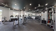 This fitness center offers a comprehensive workout environment designed to accommodate a wide range of exercise routines. The space is defined by its distinctive dark shiplap ceiling, which contrasts sharply with the bright white walls to create a modern, stylish atmosphere. Recessed lighting and multiple ceiling fans ensure the space remains well-lit and comfortable during workouts.

The cardio zone features a row of treadmills and elliptical machines positioned along large windows that invite generous natural light and outdoor views, enhancing the workout experience. A dedicated strength training area includes a full rack of dumbbells, multiple cable machines, and a cable functional trainer, providing ample options for free weight and resistance training. Several weight benches are distributed throughout the space for versatile use.

A section of the fitness center is lined with floor-to-ceiling mirrors, which amplify the sense of space and allow users to monitor their form during exercise. The flooring throughout consists of durable dark grey carpet tile with subtle accent lines, practical for a high-traffic fitness environment. Motivational artwork is displayed on the walls, adding energy to the space. Glass-paneled doors lead to an outdoor area, further connecting the interior to the surrounding environment.