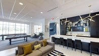 This sophisticated single-floor amenity space is designed with a contemporary aesthetic, blending functionality with upscale style. The open-concept layout connects several distinct zones, creating a seamless flow throughout the space. A striking bar area anchors one side of the room, featuring sleek dark cabinetry, a white quartz countertop, and bar stool seating. Above, a sculptural gold sputnik chandelier adds a bold focal point, complementing the dark upper shelving adorned with curated décor. Adjacent to the bar, a comfortable lounge area offers plush seating with yellow and neutral accent pillows atop a bold striped area rug, creating an inviting social atmosphere. The space extends into a game room area, which includes a billiards table and modern pod-style seating, perfect for recreation and casual gatherings. Natural light filters in through large windows, brightening the white drop-ceiling panels fitted with recessed lighting. A media area with a wall-mounted flat-screen television provides an additional entertainment option. The overall palette of white, charcoal, and gold accents gives the space a polished, modern character suitable for both relaxed and social occasions.