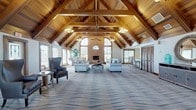 This single-floor clubhouse is defined by its dramatic architectural centerpiece: a cathedral ceiling with rich, warm-toned wood planking and exposed timber trusses that span the full length of the expansive main gathering hall. Pendant chandeliers and ceiling fans complement the natural light streaming through numerous windows and a prominent arched window above the brick fireplace, which serves as a striking focal point along the main wall. The main hall is generously proportioned, accommodating multiple seating arrangements on patterned carpet, creating distinct lounging zones suitable for both intimate conversation and larger group gatherings. The entry foyer continues the vaulted wood ceiling aesthetic, welcoming guests with a decorative console, round mirror, and a glass-paneled front door. Transitioning through a connecting passage, the game room offers a contrasting, more casual atmosphere with a polished tile floor, standard ceiling height, pendant lighting, and a full-size billiard table as its centerpiece. Additional seating and café-style tables line the perimeter, while windows provide natural light and outdoor views. Together, the spaces offer a well-rounded, inviting environment suited for a variety of community activities.