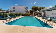 Located at 25001 SW 130th Ave in Princeton, Florida, this outdoor amenity space centers around a generously sized rectangular swimming pool with crystal-clear turquoise water, complete with stainless steel entry railings and clearly marked depth indicators. The expansive pool deck is finished in a warm beige tone, providing ample space for lounging and circulation around the pool's perimeter. Stylish teal-upholstered chaise lounges and coordinating side tables are arranged along the sun deck, accompanied by shade umbrellas that offer relief from the South Florida sun. The space is framed by neatly manicured tall hedges and tropical plantings, including palms and large potted specimens, creating a sense of privacy and lush resort-like ambiance. A covered pergola structure with a corrugated metal roof runs along one side of the pool, providing shaded access to adjacent facility rooms and amenities. The surrounding multi-story white building with modern architectural detailing forms a clean backdrop to the outdoor space. The overall design reflects a contemporary, resort-inspired aesthetic well-suited to Florida's sunny climate, offering a well-appointed and inviting environment for outdoor relaxation.