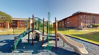 This outdoor playground is situated at 6300 Century City S in Reynoldsburg, Ohio, nestled within a well-maintained property surrounded by classic brick buildings. The play area is anchored by a multi-component play structure finished in green and brown tones, featuring platforms, climbing elements, slides, and safety railings designed to engage children of various ages. The structure rises to an accessible height, offering a variety of play experiences within a compact footprint. The ground surface beneath and around the equipment consists of blue rubber gravel, providing a cushioned, safety-conscious base. The surrounding landscape is lush and green, with well-kept grass extending across the open yard areas adjacent to the playground. Mature trees dot the property, offering natural shade and contributing to the welcoming outdoor ambiance. The brick residential buildings that frame the space feature a warm, traditional architectural style with wood accent paneling and pitched rooflines. Under open skies with ample natural light, this playground serves as an inviting recreational amenity for the surrounding community, combining functional play equipment with a safe, attractive outdoor environment.