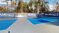 The Midland Falls Apartment Homes pickleball complex is a well-appointed outdoor recreational amenity designed for active residents. The facility comprises multiple side-by-side pickleball courts, each finished in a striking two-tone blue playing surface with crisp white boundary lines, offering excellent visibility and professional-grade playability. The courts are built on durable interlocking tile flooring in a neutral gray tone, providing a safe, all-weather surface that extends generously beyond the playing areas.

Each court is equipped with regulation nets featuring branded Midland Falls banners, adding a polished, community-branded aesthetic to the space. The complex is enclosed by a black metal fence with gated access, ensuring a secure and defined playing environment. Tall, mature pine trees line the perimeter, creating a natural screen that offers shade, privacy, and a tranquil wooded ambiance. The surrounding landscaping includes manicured shrubs and seasonal foliage, further enhancing the outdoor setting.

Abundant natural light floods the courts throughout the day, and light poles are positioned around the facility, suggesting the option for extended evening play. Views of the broader community, including the clubhouse building, are visible from the courts, reinforcing the sense of an integrated, resort-style residential amenity.