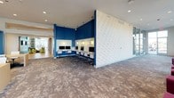 The Student Pod Lounge is a thoughtfully designed communal space that blends comfort and functionality to support student life. The lounge is anchored by soaring floor-to-ceiling windows that frame outdoor views and bathe the interior in generous natural light, creating an open and energizing atmosphere. Colorful, modular seating arrangements — featuring patterned upholstered chairs and vibrant pink ottomans — are positioned throughout the lounge area, offering flexible spaces for casual study, group conversations, or relaxation. Bold abstract artwork adorns the walls, adding a creative and lively character to the environment. A dedicated computer workstation area is defined by rich navy blue accent walls and pendant lighting overhead, providing a focused setting equipped with multiple desktop computers and ergonomic chairs for individual work. Separate semi-private study pods, set against a decorative feature wall with a graphic pattern, offer additional workspace with wooden tables and seating for small groups. Patterned carpeting ties the various zones together, while recessed lighting ensures the space remains well-lit throughout. The Student Pod Lounge is a dynamic, multi-functional environment designed to accommodate a wide range of academic and social activities.