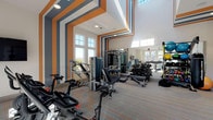 Located at 4407 Hobson Rd in Morrisville, NC, this well-appointed fitness center delivers a dynamic workout environment defined by striking architectural design and a comprehensive selection of equipment. The space is immediately distinguished by its soaring ceilings and bold graphic wall treatments featuring alternating orange, blue-gray, and white stripes that extend from floor to ceiling, creating an energizing atmosphere. Large windows along multiple walls flood the interior with natural light, offering views of the surrounding community and contributing to an open, airy feel. The flooring consists of striped, multi-toned carpet that complements the room's color palette. The equipment layout is thoughtfully organized, with a dedicated cardio zone featuring treadmills, ellipticals, and Peloton spin bikes. A strength training area houses cable machines, multi-station weight systems, and a full rack of dumbbells, while a functional training zone provides medicine balls, kettlebells, stability balls, and a heavy bag. Full-length mirrors enhance the sense of depth throughout. A wall-mounted flat-screen TV adds convenience for cardio users. Entry is provided through sleek black-framed glass doors, connecting the fitness center to the broader facility.