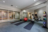 Located at 8610 N Kansas Ave in Kansas City, Missouri, this single-floor fitness center is thoughtfully designed to support a wide range of workout routines. The space is divided into distinct functional zones, each tailored to a specific type of exercise. The main studio area features a carpeted floor laid with multiple exercise mats and stability balls in various sizes, providing an ideal environment for yoga, stretching, and core training. A wall-mounted television and a rack of color-coded dumbbells are positioned along one wall, accompanied by TRX suspension straps for functional training. Recessed lighting throughout keeps the space well-lit, while neutral gray and blue tones on the walls contribute to a calm, focused atmosphere. An adjacent cycling area houses stationary bikes equipped with individual screens, allowing for interactive or guided ride sessions. Natural light filters in through windows that offer views of the surrounding outdoor area. Glass sliding doors connect the studio zones, creating an open and airy feel while maintaining visual separation between workout areas. A restroom is also accessible from within the facility. The overall design is modern and clean, prioritizing both functionality and comfort for a well-rounded fitness experience.