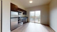 This single-floor unit presents a clean, modern aesthetic throughout its open layout. The kitchen is well-appointed with a full suite of stainless steel appliances, including a refrigerator, electric range, over-the-range microwave, and dishwasher. Rich, dark espresso-toned cabinetry provides ample upper and lower storage, while granite countertops add a polished, durable work surface. The kitchen flows naturally into the living area, which is anchored by light-toned hardwood-style flooring that extends throughout the unit, creating a cohesive and airy feel.

A standout feature of the living space is a full-width sliding glass door that opens to a private balcony, allowing natural light to fill the interior and offering views of the surrounding neighborhood and distant hills. The neutral, warm wall tones complement the flooring and enhance the brightness of the space.

The bathroom features the same light hardwood-style flooring and is accessed through a paneled white door, maintaining a consistent design language. A separate closet and utility panel are also present, ensuring practical storage and mechanical access. Overall, the unit delivers a contemporary, move-in-ready environment with thoughtful finishes and functional design.