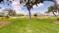 Located at 11300 Pinckney Marsh Lane in Port Royal, South Carolina, this outdoor common area offers a thoughtfully landscaped environment that reflects the natural character of the South Carolina Lowcountry. The grounds are defined by expansive, well-kept lawns that provide generous open space for recreation and relaxation. Towering live oak trees, draped in iconic Spanish moss, serve as the visual anchors of the space, casting broad canopies of shade across the lawn and contributing to the area's distinctly coastal Southern atmosphere.

Hammocks are strung between trees near the residential buildings, offering casual lounging options in a shaded setting. Adirondack-style seating is positioned throughout the grounds, taking advantage of the natural landscape and allowing residents to enjoy the serene surroundings. Native plantings, including ornamental grasses and subtropical foliage, are interspersed among the trees, adding texture and visual interest while complementing the natural environment.

The grounds open toward a treeline that hints at nearby marsh and water views, consistent with the property's location along Port Royal's scenic waterfront corridor. The multi-story residential buildings visible in the background provide context for the scale of the community. Overall, the outdoor space conveys a calm, nature-integrated environment well-suited to the Lowcountry setting.
