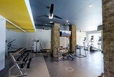 This fitness center combines industrial design with modern functionality. The space is defined by striking exposed brick columns that rise to a dark-painted ceiling, creating a bold contrast against the light gray walls. Ceiling fans are strategically placed throughout, ensuring comfortable airflow during workouts. Recessed and track lighting illuminate the floor, which is finished in a durable, polished dark concrete suitable for high-traffic use. A flat-screen TV is mounted between the brick columns, offering entertainment while exercising. The strength training area is well-equipped with a cable machine, adjustable bench, and a dedicated free weights section featuring a dumbbell rack. On the opposite side, the cardio zone houses elliptical machines and additional equipment, catering to a range of fitness routines. A tall, narrow window along one wall introduces a touch of natural light and a pop of yellow, adding visual energy to the space. The overall ambiance is urban and contemporary, balancing an industrial aesthetic with a well-organized, fully functional workout environment.