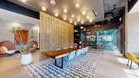 Located at 307 SW 5th St in Fort Lauderdale, this single-floor commercial space offers a thoughtfully designed interior that blends modern aesthetics with bold, eclectic style. The main open area is flooded with natural light through a full-length floor-to-ceiling glass window wall, revealing lush greenery beyond and creating a bright, welcoming atmosphere. A continuous orange banquette runs along the windowed facade, paired with vibrant yellow chairs and pendant globe lighting overhead, establishing a stylish lounge and casual seating zone. A large live-edge communal table surrounded by light blue chairs anchors the central dining or collaboration area, set atop a graphic black-and-white script-patterned rug. A striking floor-to-ceiling slatted wood screen divides the space, serving as both a design statement and functional partition. An open metal shelving unit, adorned with curated décor objects, adds visual depth and warmth. Exposed brick columns and dark painted ceilings contribute an industrial edge, while vibrant red accent chairs and bold artwork inject color and personality. Glass-enclosed meeting rooms with teal walls, coral chairs, and light wood benches provide private workspaces. Together, these elements create a dynamic, energetic environment ideal for collaboration and community.