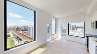 This contemporary one-bedroom, one-bathroom residence offers a well-planned layout across a single floor, thoughtfully designed to maximize both comfort and functionality. Light hardwood flooring flows seamlessly throughout the entire unit, lending warmth and visual continuity to the modern interior.

The open living and kitchen area is a standout, anchored by large black-framed windows that flood the space with natural light and frame expansive urban skyline views. The kitchen is outfitted with full stainless steel appliances — including a range, microwave, and refrigerator — complemented by sleek white cabinetry, clean countertops, and a striking blue hexagonal tile backsplash that adds a bold design accent. A wall-mounted mini-split HVAC unit provides efficient climate control.

The bedroom is a bright and serene retreat, featuring corner windows with roller shades that offer both natural light and privacy, along with its own wall-mounted HVAC unit. The hallway connects the rooms efficiently, with recessed lighting and clean white walls maintaining the unit's crisp, modern aesthetic.

The bathroom is appointed with a full soaking tub, a tiled surround featuring a playful mix of white and teal tiles, and a white vanity with a clean countertop. Natural light enters through a window above the tub, enhancing the fresh, airy feel of the space.