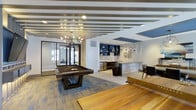 This single-floor entertainment space is thoughtfully designed to serve as a dynamic social hub. The room immediately draws attention with its striking ceiling treatment, featuring alternating striped panels that add visual depth and a contemporary architectural character. Recessed lighting and a statement chandelier with a starburst silhouette provide warm, layered illumination throughout.

At the center of the space sits a substantial pool table, positioned atop mixed-tone hardwood flooring that adds warmth and texture underfoot. A large wooden surface—likely a game or community table—occupies the foreground, reinforcing the room's recreational focus.

The wet bar area along the back wall is a standout feature, finished with a textured stone or concrete-style counter and flanked by bold blue open shelving stocked for entertaining. A stainless steel beverage refrigerator is integrated into the cabinetry, and pendant lighting accents the bar zone. Decorative wall sculpture nearby adds an artistic touch.

Beyond a glass-paneled door, a glimpse of fitness equipment suggests an adjacent exercise room, enhancing the overall amenity offering. The space balances modern aesthetics with functional design.