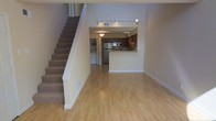 This two-story residence offers a well-designed open-concept main level that seamlessly connects the living area and kitchen. The main floor features light hardwood flooring that extends throughout, creating a bright and cohesive aesthetic. Elevated ceilings contribute to a sense of spaciousness, while a track lighting system installed along an overhead rail provides ample illumination throughout the living area.

The kitchen is positioned at the rear of the main level and is accessible via a breakfast bar counter that serves as a natural divider between the cooking and living spaces. Dark wood cabinetry lines the kitchen walls, complemented by stainless steel appliances, including a refrigerator and microwave. A pendant light fixture adds a finishing touch above the kitchen area. A dedicated laundry area is also visible adjacent to the kitchen, offering added convenience.

A carpeted staircase runs along one wall of the main level, leading to the upper floor. Neutral, warm-toned walls throughout complement the flooring, giving the space an inviting and functional character overall.