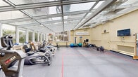 The Atrium Fitness Center at 551 Gibson Ave in Pacific Grove, California, is a thoughtfully designed fitness facility that stands out for its remarkable glass-paneled atrium ceiling, which floods the entire space with natural light and creates a bright, energizing atmosphere throughout the day. The soaring transparent roof is supported by a clean framework of structural beams, lending the space an open, greenhouse-like character that feels welcoming and expansive.

The main exercise floor is finished with durable grey interlocking mat tiles marked with colored lines that define training zones, providing both functionality and visual organization. Large windows along the perimeter walls frame views of surrounding greenery, reinforcing the connection to the outdoors. Ceiling fans are mounted overhead to ensure comfortable airflow during workouts.

The facility is well-equipped with a wide range of fitness equipment, including cardio machines such as a treadmill, elliptical, and rowing machine, as well as strength-training equipment including a cable machine, weight bench, and stability balls. A wall-mounted ballet barre and a stall bar add versatility for flexibility and rehabilitation exercises. A wall-mounted flat-screen television, a clock, and wall-mounted speakers enhance the overall experience. A small administrative area with filing cabinets and a water cooler is tucked near the entrance, completing this comprehensive and inviting fitness environment.