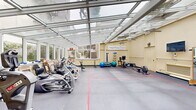 The Atrium Fitness Center at 551 Gibson Ave in Pacific Grove, California, is a thoughtfully designed fitness facility that stands out for its remarkable glass-paneled atrium ceiling, which floods the entire space with natural light and creates a bright, energizing atmosphere throughout the day. The soaring transparent roof is supported by a clean framework of structural beams, lending the space an open, greenhouse-like character that feels welcoming and expansive.

The main exercise floor is finished with durable grey interlocking mat tiles marked with colored lines that define training zones, providing both functionality and visual organization. Large windows along the perimeter walls frame views of surrounding greenery, reinforcing the connection to the outdoors. Ceiling fans are mounted overhead to ensure comfortable airflow during workouts.

The facility is well-equipped with a wide range of fitness equipment, including cardio machines such as a treadmill, elliptical, and rowing machine, as well as strength-training equipment including a cable machine, weight bench, and stability balls. A wall-mounted ballet barre and a stall bar add versatility for flexibility and rehabilitation exercises. A wall-mounted flat-screen television, a clock, and wall-mounted speakers enhance the overall experience. A small administrative area with filing cabinets and a water cooler is tucked near the entrance, completing this comprehensive and inviting fitness environment.