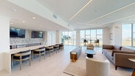 Located at 7000 Kiran Patel Dr in Winter Garden, Florida, this sophisticated single-floor lounge and living space offers a refined, contemporary environment designed for comfort and social engagement. The open-plan layout is anchored by large-format light tile flooring that flows seamlessly throughout, lending a clean and cohesive aesthetic to the entire space.

Floor-to-ceiling window walls line the perimeter, flooding the interior with abundant natural light and framing sweeping views of a nearby waterway and the surrounding Florida landscape. The living area features multiple seating arrangements, including plush sofas, curved accent chairs, and high-top bistro tables, all styled with a neutral palette accented by gold metallic details.

A built-in bar and dining counter with wood-toned cabinetry and bar seating serves as a central gathering point, complemented by dual wall-mounted televisions and a striking textured mosaic backsplash. Linear LED lighting recessed into tiered ceiling details adds a modern architectural touch and enhances the sense of depth and volume.

Intimate conversation nooks are thoughtfully placed near the windows, offering quiet spots to relax while taking in the outdoor scenery. The overall ambiance is bright, airy, and polished — reflecting a well-designed communal environment that balances openness with warmth.