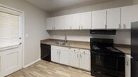 This single-story residence presents a straightforward, functional layout with a neutral color palette throughout. The living area is finished with wall-to-wall carpet in a warm beige tone, complemented by soft gray walls and clean white trim. A window with blinds allows natural light to brighten the room, and a front entry door provides direct access to the space.

The kitchen is equipped with white cabinetry — both upper and lower — offering ample storage. A double-basin stainless steel sink is set into laminate countertops, flanked by black appliances including a dishwasher and an electric range. Wood-look vinyl flooring runs throughout the kitchen, providing a durable, easy-to-clean surface.

The bedrooms are carpeted and painted in the same neutral tones seen throughout the unit. Each room features windows with blinds that bring in natural light and offer outdoor views of surrounding greenery. One bedroom includes a ceiling light fixture, and closet access is visible nearby.

The bathroom features a tiled tub-shower combination, a toilet, and a vanity, with wood-look vinyl flooring matching the kitchen. Louvered closet doors are visible at the entry to the bathroom, adding a classic detail. Overall, the space presents a clean, livable environment with consistent finishes.