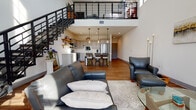 Located at 200 W Grand Ave in Beloit, Wisconsin, this two-story loft showcases a thoughtfully designed open-concept layout that blends modern aesthetics with functional living. The main floor is anchored by a dramatic double-height ceiling that creates a striking sense of volume and openness. Warm hardwood flooring runs throughout, complementing the clean white walls and contemporary finishes.

The living area is generously proportioned, with large windows that invite abundant natural light and offer views of the surrounding streetscape. A bold industrial-style staircase with sleek black steel railings serves as both a functional and design centerpiece, connecting the two floors.

The kitchen features gray shaker cabinetry, white quartz countertops, stainless steel appliances including a French-door refrigerator and dishwasher, and a central island with bar seating — ideal for both casual dining and entertaining.

A well-appointed full bathroom on the main level includes a gray vanity with quartz countertops and polished chrome fixtures. Additional space on the main floor accommodates a dedicated office or flex area filled with natural light.

The upper level opens to a loft-style bedroom with plush carpeting, multiple windows, and a second full bathroom. The industrial railing along the upper landing reinforces the loft's contemporary character while maintaining open sightlines to the floor below.