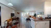 This single-floor residence is designed with a clean, contemporary aesthetic, characterized by crisp white walls, light wood-style flooring, and an abundance of natural light throughout. A hallway entrance leads into the heart of the home, where the living, dining, and kitchen areas flow together in an open-plan layout.

The living room serves as the central gathering space, featuring a sliding glass door that opens directly onto a private balcony with outdoor seating and tree-lined views. A second window fitted with white plantation shutters adds further natural light. The space transitions seamlessly into a defined dining area, which accommodates a round glass-top table and benefits from the same open, airy atmosphere.

The galley-style kitchen is efficiently laid out with white shaker cabinetry, dark countertops, and a full suite of stainless steel appliances including a refrigerator, range, microwave, and dishwasher. A peninsula counter connects the kitchen to the living and dining areas.

The bedroom is a calm, well-proportioned retreat with a ceiling fan, a window with plantation shutters offering outdoor views, and access to a walk-through closet. The bathroom features a tiled soaking tub and shower combination with a clean, minimalist finish and marble-look flooring.