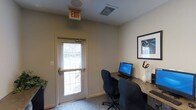 This single-floor space functions as a small yet practical business center designed to support productivity and convenience. The room features a built-in desk along the wall, equipped with a desktop computer and keyboard, providing a functional workstation setup. Two ergonomic office chairs offer comfortable seating for users working side by side. The space is illuminated by a combination of overhead recessed lighting and natural light filtering through a windowed exterior door fitted with horizontal blinds, which also provides direct access to the outdoors. The neutral-toned walls and light carpeted flooring contribute to a clean, professional atmosphere. A framed piece of wall art adds a modest decorative touch to the otherwise utilitarian environment. An exit sign and HVAC vent are visible overhead, reflecting the space's compliance with safety and comfort standards. The overall layout is efficient and straightforward, making it well-suited as a shared amenity space for tasks such as printing, browsing, or general computer use.