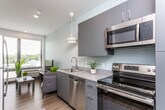 This contemporary single-floor unit offers a cohesive and functional layout with modern finishes throughout. The open-concept living and kitchen area creates an inviting, connected atmosphere. The kitchen is well-equipped with stainless steel appliances — including a range, dishwasher, microwave, and refrigerator — complemented by sleek gray cabinetry, light countertops, a glass tile backsplash, and a stylish pendant light. The living area features a comfortable seating arrangement centered around a media console, with large windows fitted with horizontal blinds that allow ample natural light and provide outdoor views of the surrounding neighborhood. Warm hardwood-style flooring flows seamlessly throughout the space, adding a sense of continuity and warmth. The bedroom is a calm, restful retreat with a large window that brings in natural light, clean white walls, and rich wood flooring. A dedicated closet space with built-in shelving offers practical storage. The bathroom is clean and modern, featuring a soaking tub with shower, a gray floating vanity with white countertops, and the same warm wood-tone flooring. The overall aesthetic is minimalist yet comfortable, with neutral tones and contemporary fixtures creating a polished, move-in-ready environment.