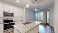 Located at 10387 Vista Oaks Ct in Orlando, FL, this well-appointed one-bedroom, one-bathroom residence offers a thoughtfully designed open-concept floor plan. The combined kitchen and living area serves as the heart of the home, highlighted by rich wood-style flooring, recessed lighting, and a ceiling fan that enhances airflow and comfort. The kitchen is a standout feature, showcasing white shaker-style cabinetry extending to the ceiling, a classic subway tile backsplash, granite countertops, and a central island with a double stainless steel sink and pendant lighting overhead. Stainless steel appliances, including a range, microwave, and refrigerator, complete this modern culinary space. The living area flows naturally toward a large window and glass door that opens to a private balcony, allowing ample natural light throughout. The bathroom is clean and contemporary, featuring a granite-topped vanity with white cabinetry, wood-style flooring, and a tiled walk-in shower with glass sliding doors. The bedroom offers plush carpeting, a ceiling fan, and two closets for generous storage. Additional conveniences include an in-unit laundry area. The overall aesthetic is bright, modern, and move-in ready.