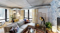 This single-floor urban loft blends industrial architecture with vibrant, eclectic interior design. Exposed concrete ceilings and visible metallic ductwork run throughout the space, lending a raw, loft-style character that is complemented by warm, colorful decor choices. Large floor-to-ceiling windows frame urban cityscape views in multiple rooms, flooding the interiors with natural light.

The living area features a comfortable sectional sofa anchored by a patterned area rug, with bold yellow geometric curtains and an array of curated accents creating a lively, layered atmosphere. A separate sitting nook offers a cozy retreat, dressed in floral wallpaper, botanical curtains, and soft seating with richly textured throw pillows.

The kitchen is well-appointed with stainless steel appliances, including a refrigerator, electric range, and dishwasher, complemented by granite countertops, a mosaic tile backsplash, sleek cabinetry, and track lighting overhead. A corner reading or lounge area provides additional flexibility, bathed in light from wraparound windows.

The bedroom offers direct city views through a large window, with exposed concrete and ductwork continuing the industrial aesthetic. The bathroom makes a bold statement with wide blue-and-white horizontal stripes, herringbone tile flooring, a combined tub and shower, and a clean white vanity. A second bathroom features a glass-enclosed shower and connects seamlessly to the bedroom.