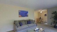 This single-floor residence offers a thoughtfully arranged open-plan layout that connects the living and dining areas in a cohesive, functional design. The living area is anchored by a plush gray sofa accented with decorative pillows in navy blue and patterned tones, complemented by a glass-topped coffee table that adds a touch of lightness to the space. A large canvas print depicting a dramatic landscape adds a vibrant focal point to the soft, neutral-toned walls, enhancing the room's visual character. Natural light filters through the space, contributing to a bright and welcoming ambiance. The dining area flows seamlessly from the living room and features a wooden table with matching chairs, ideal for everyday dining. A ceiling fan with a built-in light fixture provides both comfort and illumination to the dining zone. The consistent use of light wall tones and neutral flooring throughout creates a cohesive and airy feel across the entire floor. The overall design emphasizes practicality and ease of living, making this a well-organized and comfortable single-level space suited for everyday residential use.